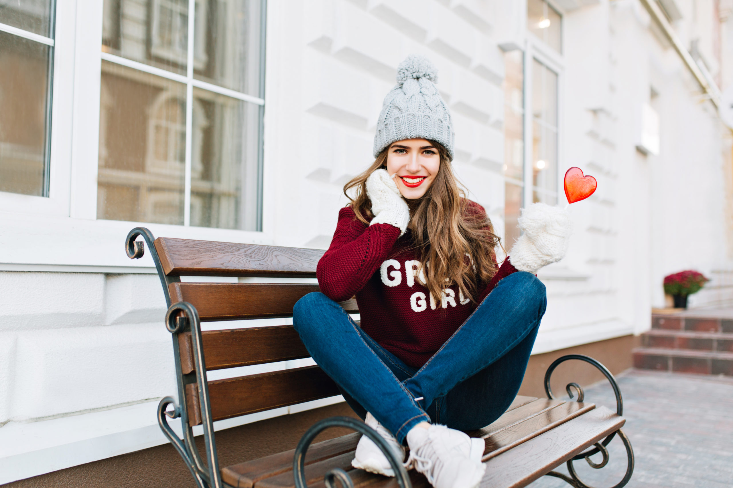 Full-lenth beautiful young girl with long hair in knitted hat and white gloves sitting on bench in city. She holds caramel heart, smiling to camera. Custom hoodie designs by OM Clothing Boston
